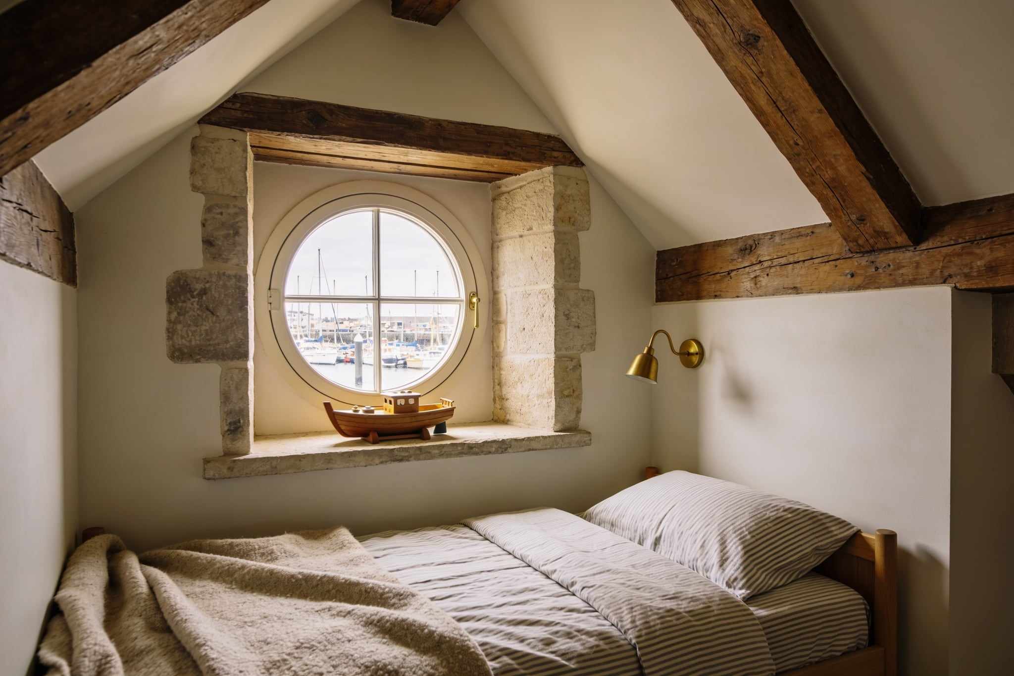 A small bed with striped linen in a nook under the eaves, a wooden toy boat on the sill of a round window