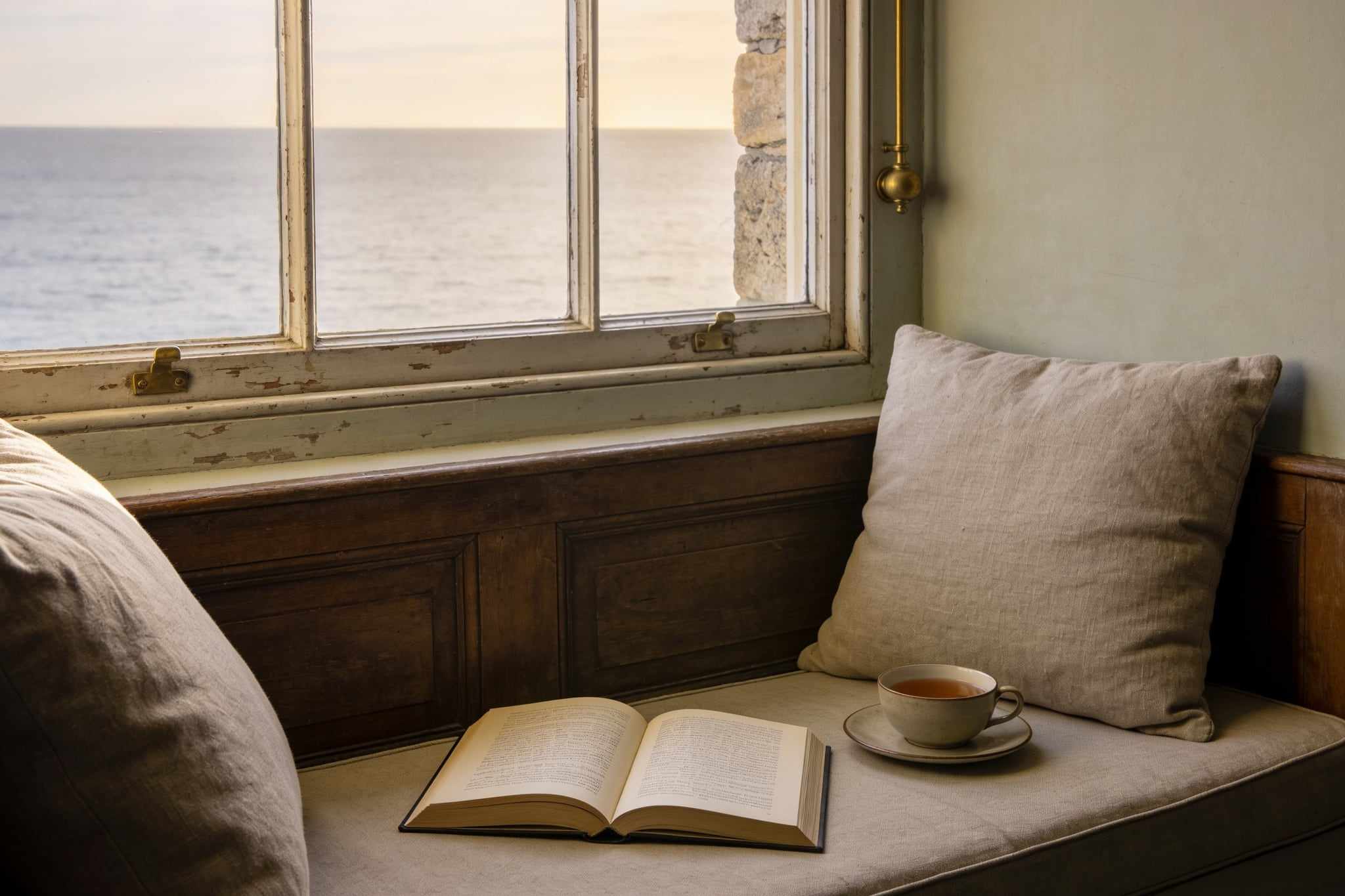 A window seat with linen cushions, an open book and a cup of tea, the sea beyond the old sash window