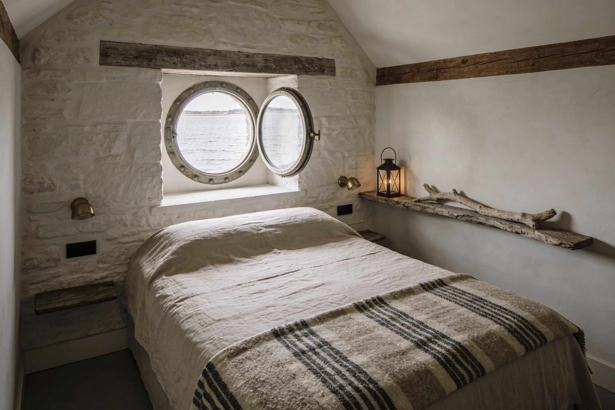 A bedroom with whitewashed stone walls, a bed with a striped wool blanket, round windows and a lantern on a driftwood shelf