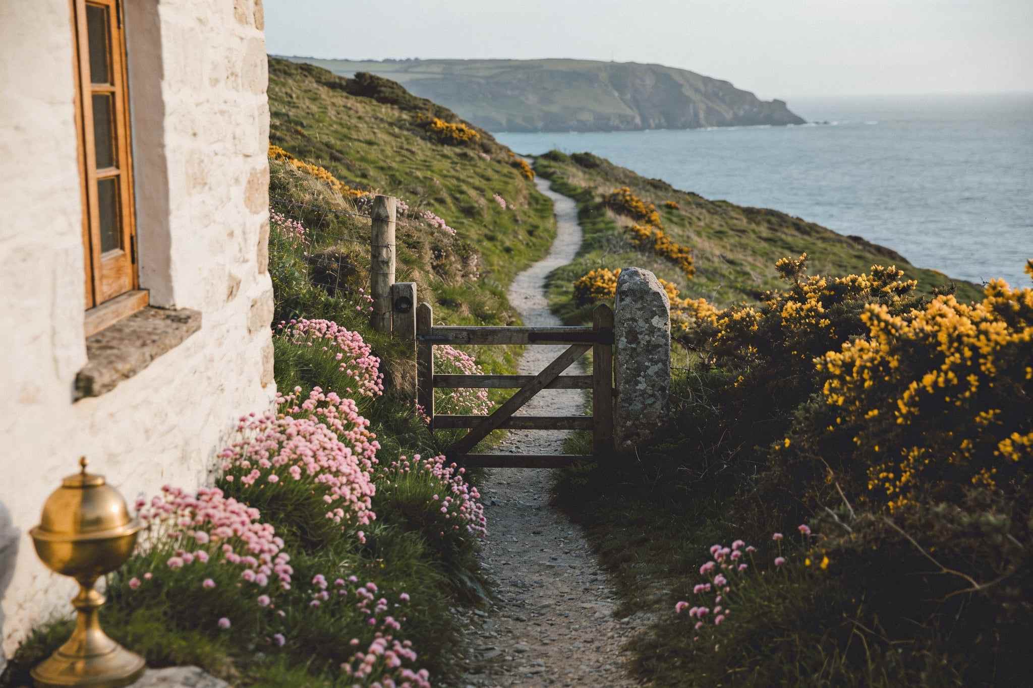 A narrow path winding along green cliff tops through sea pinks and gorse, a wooden gate and the sea beyond