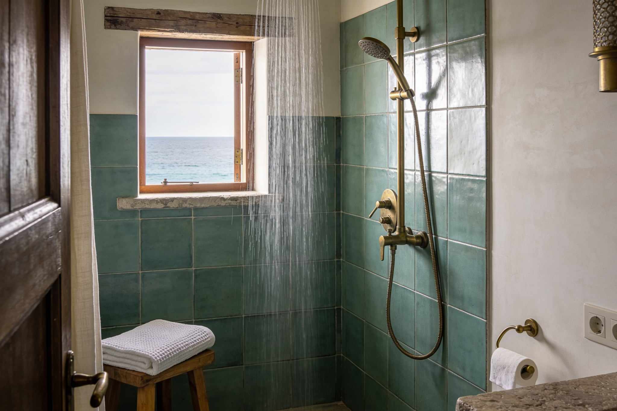 A walk-in shower with sea-green glazed tiles, a brass rain shower and a small window onto the sea
